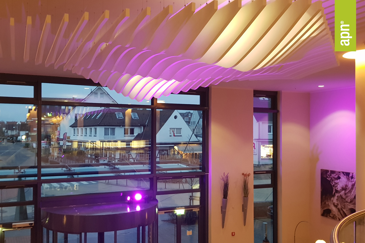 Innenansicht einer modernen Hotellobby mit großen Fenstern und Blick auf die Straße. Die Decke ist mit weißen, wellenförmigen Akustikbaffeln gestaltet, die von violettem Licht angestrahlt werden. Links unten befindet sich eine Drehtür, rechts sind dekorative Vasen mit hohen Pflanzen an der Wand platziert. Ein grünes, vertikales Banner mit der Aufschrift „apn“ ist am rechten oberen Bildrand zu sehen.