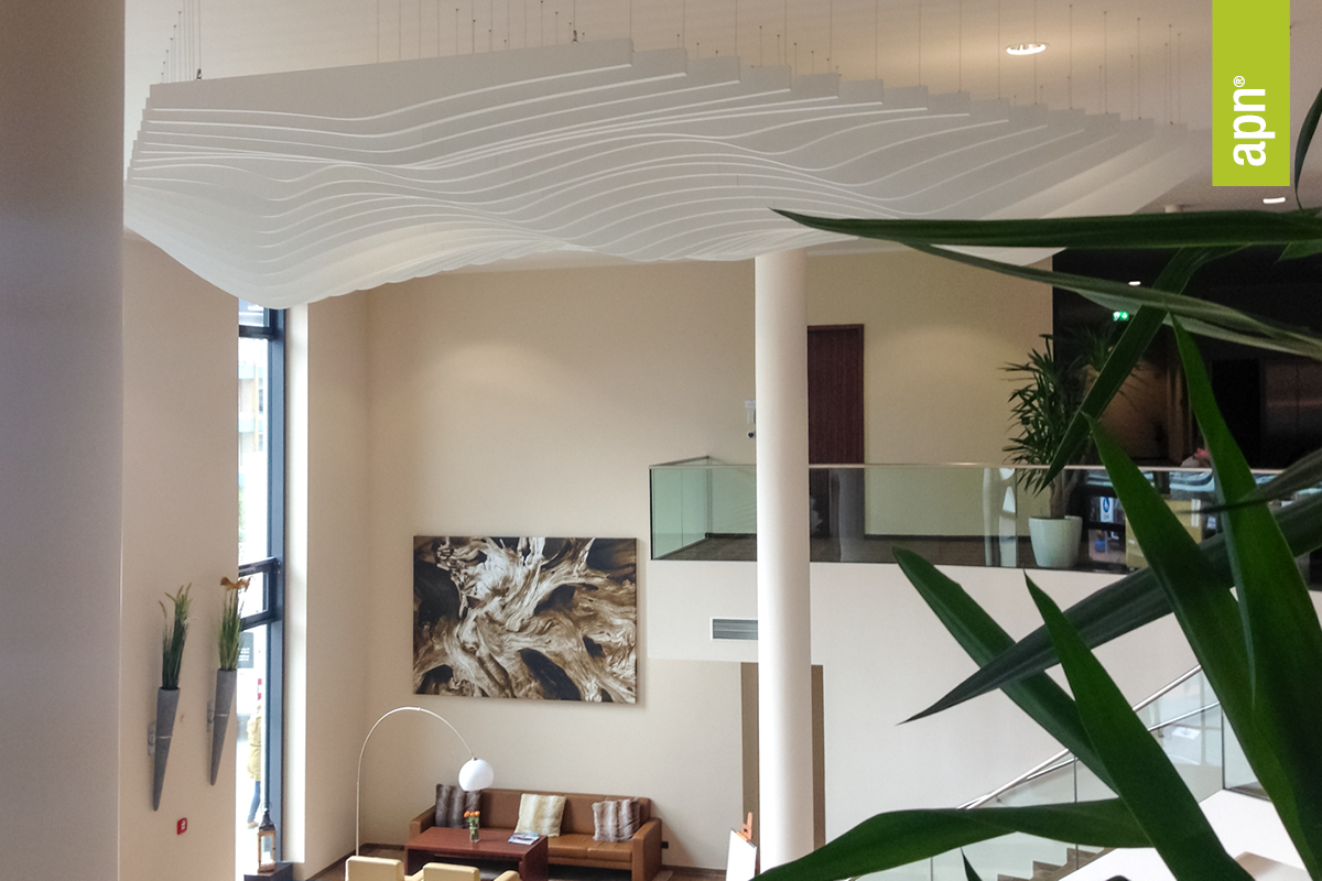 Moderne Hotellobby mit hoher Decke und wellenförmiger, weißer akustischer Deckengestaltung. Große Fenster lassen viel Tageslicht herein, Pflanzen und abstrakte Kunst an der Wand sorgen für dekorative Akzente. Im unteren Bereich befindet sich eine gemütliche Sitzecke mit Sofas und Kissen, darüber verläuft eine Galerie mit Glasgeländer. Rechts im Bild ist eine Treppe zu sehen, oben rechts ein grünes Banner mit der Aufschrift „apn“.