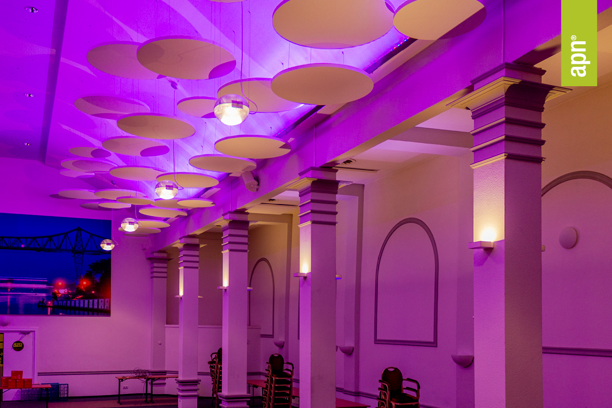 Innenraum mit mehreren Säulen und runden akustischen Deckenelementen, die in violettem Licht erstrahlen. Hängende Leuchten sorgen für zusätzliche Beleuchtung. Im Hintergrund ist ein großes Akustik-Wandbild mit einer nächtlichen Stadtansicht samt Brücke zu sehen. Es scheint sich bei der Aufnahme um einen Veranstaltungssaal eines Hotels zu handeln. Rechts im Bild befindet sich ein grünes, vertikales Banner mit der Aufschrift „apn“.