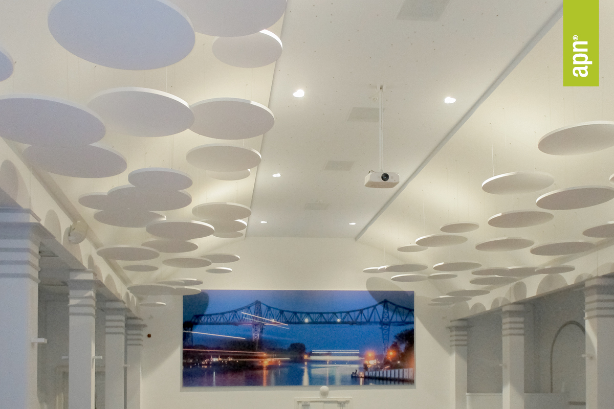 Innenraum mit hoher Decke und zahlreichen runden Akustikpaneelen, die von der Decke abgehängt sind. An der Wand hängt ein großformatiges Akustikbild einer beleuchteten Brücke bei Nacht. Der Raum ist von architektonischen Säulen gesäumt, und an der Decke ist ein Projektor montiert. Es scheint sich bei der Aufnahme um einen Veranstaltungssaal eines Hotels zu handeln. Rechts im Bild befindet sich ein grünes, vertikales Banner mit der Aufschrift „apn®“.