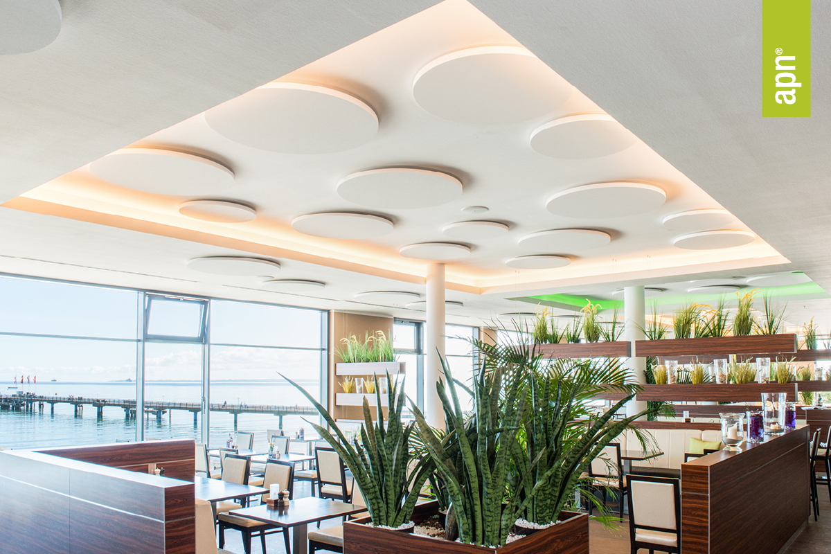Heller, moderner Restaurantinnenraum mit Blick auf das Meer und einen Steg. Die Decke ist mit runden Einbauleuchten ausgestattet, während grüne Pflanzen und Holzmöbel mit weißen Polstern eine einladende Atmosphäre schaffen. Das Bild vermittelt eine ruhige Umgebung, in der akustische Lösungen zur Klangoptimierung in der Gastronomie eine zentrale Rolle spielen. Rechts im Bild ist ein grünes Banner mit dem Schriftzug „apn“ zu sehen.
