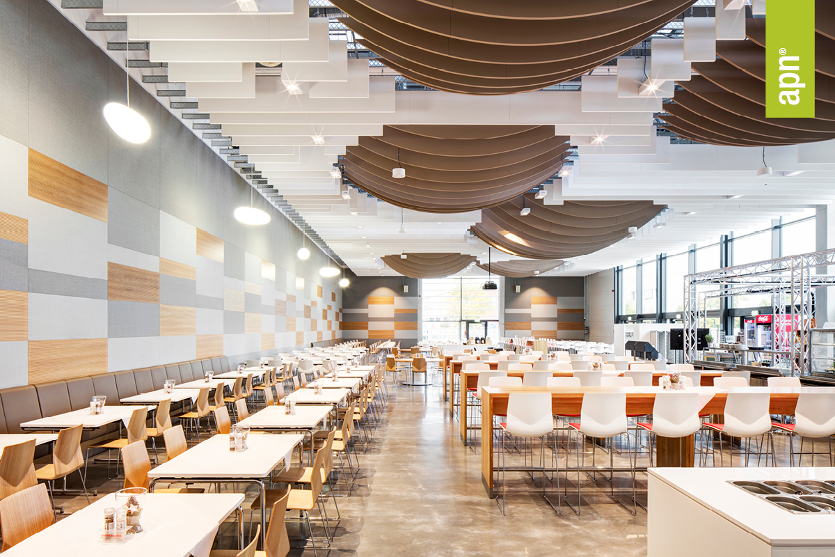 Großzügige, moderne Cafeteria mit hoher Decke und großen Fenstern auf der rechten Seite. An der Decke hängen wellenförmige, braune Paneele in Reihen. Die Wände sind mit einem Muster aus grauen, weißen und holzfarbenen Paneelen gestaltet. Im Raum stehen mehrere Reihen von Tischen und Stühlen: links lange Tische mit Holzstühlen, in der Mitte und rechts höhere Tische mit weißen Barhockern. Der Boden besteht aus poliertem Beton. Oben rechts im Bild ist ein grünes, vertikales Banner mit der Aufschrift „apn“ zu sehen.