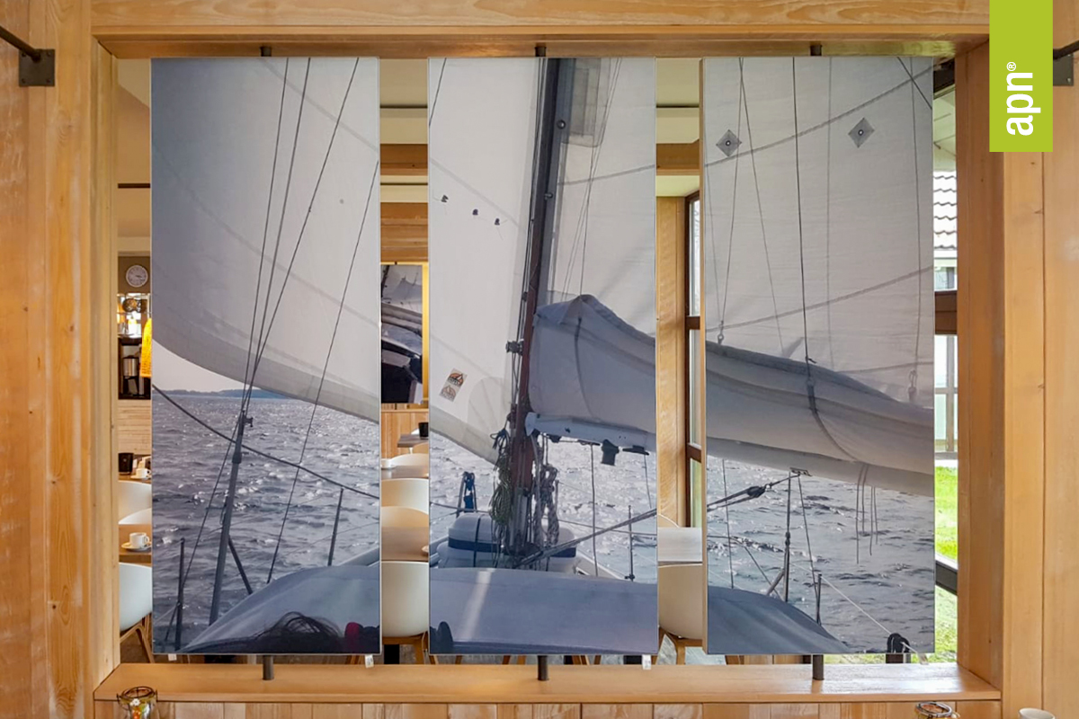 Drei vertikale Wandpaneele zeigen ein durchgehendes Fotomotiv eines Segelboots auf dem Wasser. Die Aufnahme stammt vom Deck des Boots und zeigt Segel, Takelage sowie den Blick auf Wasser und Himmel. Die Paneele sind in einem Innenraum mit Holzmöbeln und -rahmen montiert.