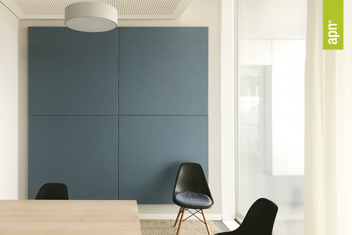 Ein moderner, minimalistisch eingerichteter Raum mit großen, quadratischen Akustik Wandabsorbern in mattem Blau. Vor der Wand stehen drei schwarze Stühle mit Holzbeinen, einer davon leicht gepolstert. Links im Bild ist ein heller Holztisch zu sehen. Die Decke hat eine weiße Lampe und ein perforiertes Deckenelement. Rechts im Bild befindet sich ein großes Fenster mit weißen Vorhängen, das viel natürliches Licht hereinlässt. Oben rechts ist das Logo "apn" in weißer Schrift auf grünem Hintergrund zu sehen.