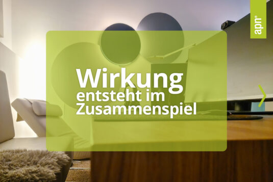 Moderner Lounge- oder Bürobereich mit weißem Sessel, Holztisch und flauschigem Teppich. Im Hintergrund befinden sich runde Wanddekorationen und eine Akustikleuchte. Auf dem Bild steht der Text „Wirkung entsteht im Zusammenspiel“. Oben rechts ist ein grünes, vertikales Banner mit der Aufschrift „apn“ zu sehen.