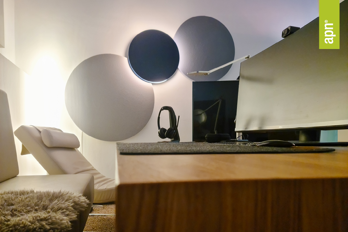Modern gestalteter, heller Raum mit weißem Loungesessel und Kissen, drei zwei Wandabsorber und eine runde Akustikleuchte in Grau- und Blautönen sowie einem Schreibtisch im Vordergrund. Auf dem Tisch stehen ein Kopfhörerständer mit Kopfhörern, ein großer Monitor und eine verstellbare Schreibtischlampe. Oben rechts im Bild befindet sich ein grünes Label mit der Aufschrift „apn“.