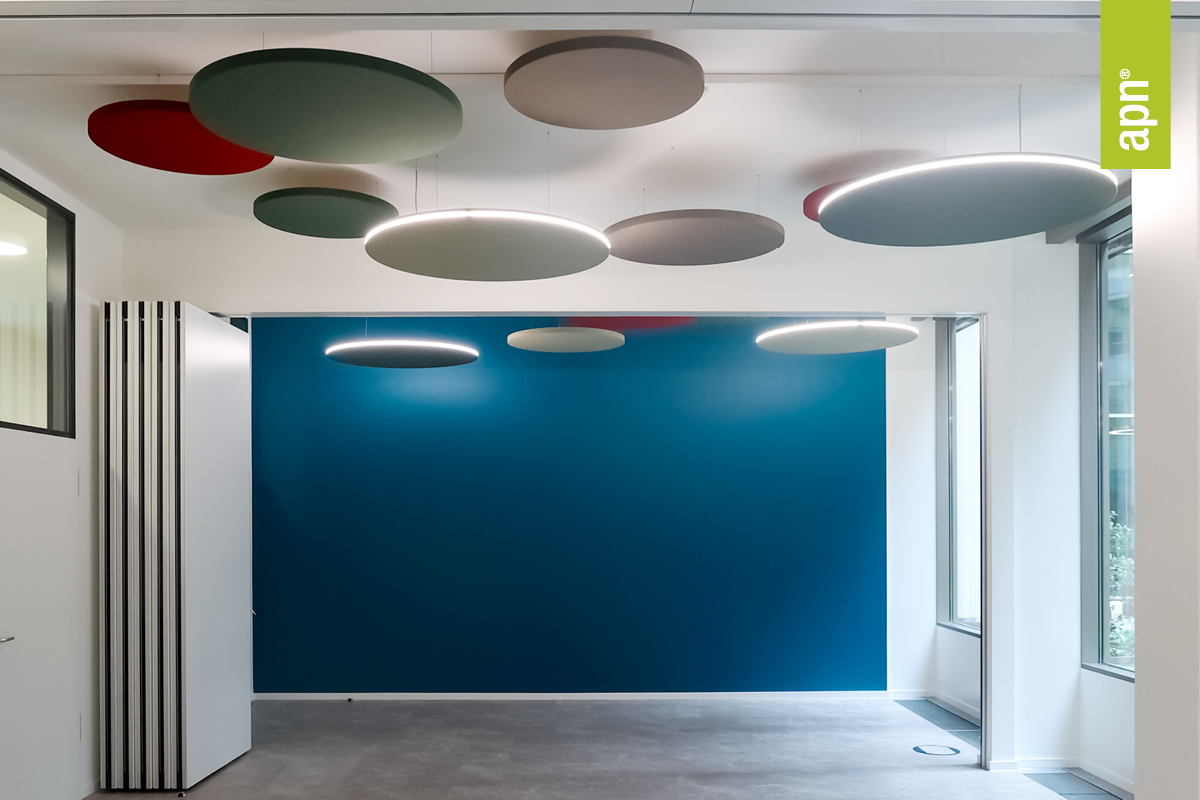 Moderner Innenraum mit blauer Akzentwand und mehreren runden Akustik-Deckenelementen in Grün, Rot und Beige, teils beleuchtet. Rechts sorgt ein Fenster für Tageslicht, links befindet sich eine Glaswand mit möglicher Trennwand. Die Raum Wirkung ist minimalistisch gestaltet mit grauem Boden. Oben rechts im Bild ist ein grünes, vertikales Banner mit der Aufschrift „apn®“ zu sehen.