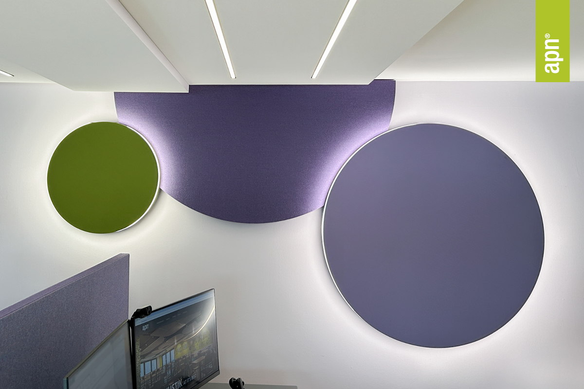Moderner Innenraum mit zwei runden Akustik-Designleuchten und einem halbkreisförmigen Wandpaneel in unterschiedlichen Größen und Farben: grün, violett (halbkreisförmig) und hellviolett. Unten links ist ein Teil eines Computerbildschirms zu sehen, an der Decke befinden sich weiter Akustikleuchten. Rechts im Bild steht vertikal das Wort „apn“.