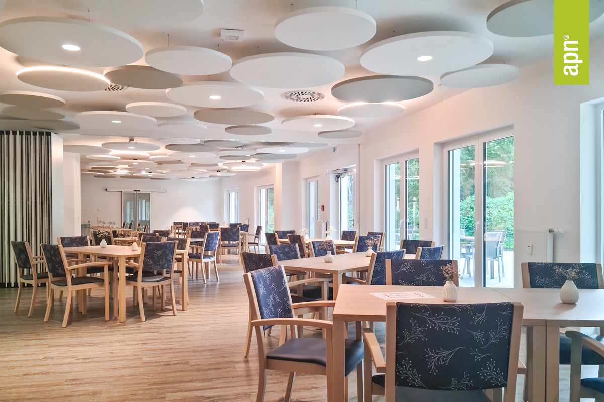 Heller, großzügiger Speiseraum mit mehreren Holztischen und Stühlen, deren Sitzflächen dunkelblaue Polster mit weißem Blumenmuster zieren. Die Decke ist mit zahlreichen runden Akustikpaneelen in Weiß- und Grautönen gestaltet. Zudem sind an der Decke weiter Akustikleuchten (rund) angebracht. Große Fenster und Glastüren auf der rechten Seite lassen viel Tageslicht herein. Der Boden besteht aus hellem Holz und unterstreicht die freundliche Wirkung des Raumes.