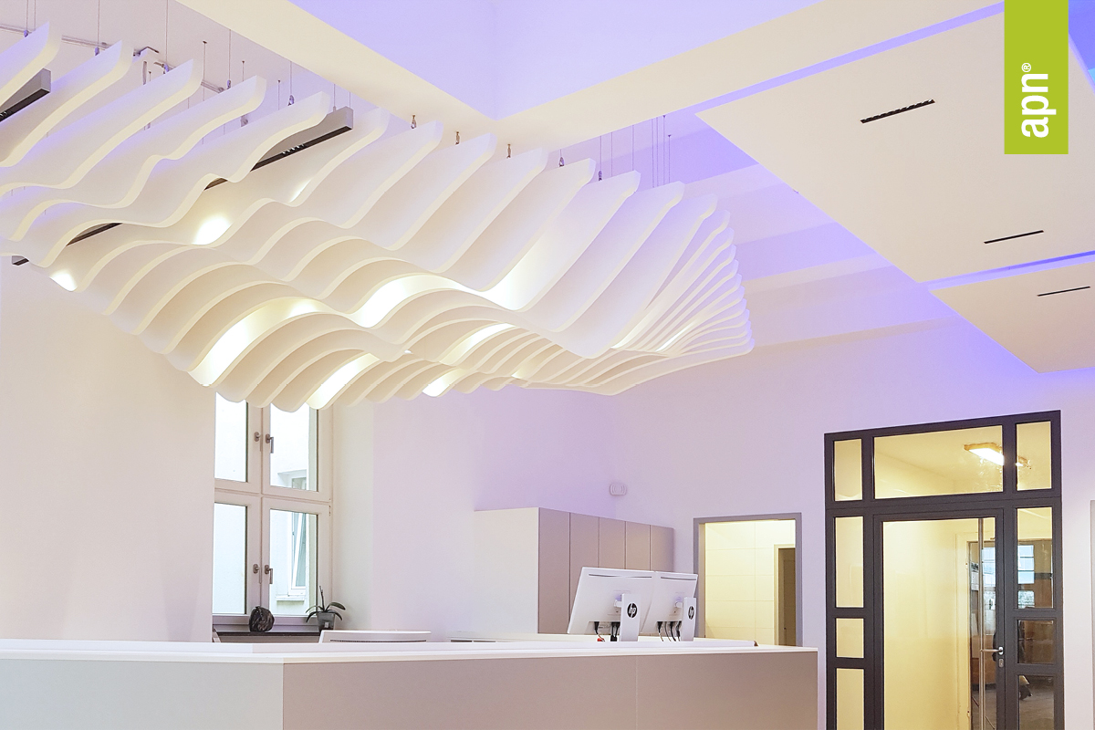 Moderner Innenraum mit wellenförmigen weißen Akustikpaneelen an der Decke zur Verbesserung der Raumakustik. Darunter befindet sich ein Empfangstresen mit zwei Monitoren. Große Fenster lassen natürliches Licht herein, und die Wände sind weiß gestrichen. Rechts ist eine Tür mit Glaspaneelen und ein grüner Banner mit 'apn'-Text.