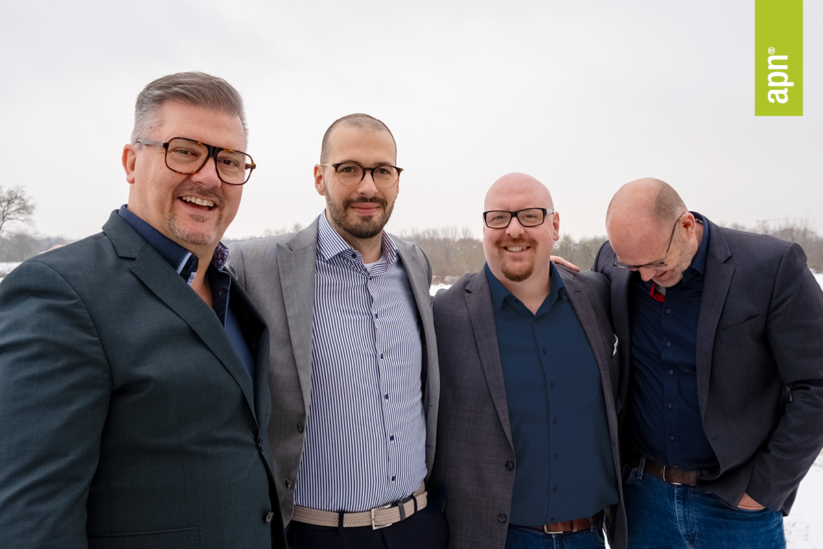 Vier Männer, alle Brillenträger, schauen in die Kamera. Alle Tragen Hemd, Jeans und Sakko. Im Hintergrund ist eine Schnee-Landschaft zu erkennen. Die Männer stehen nebeneinander und halten sich an den Schultern fest.