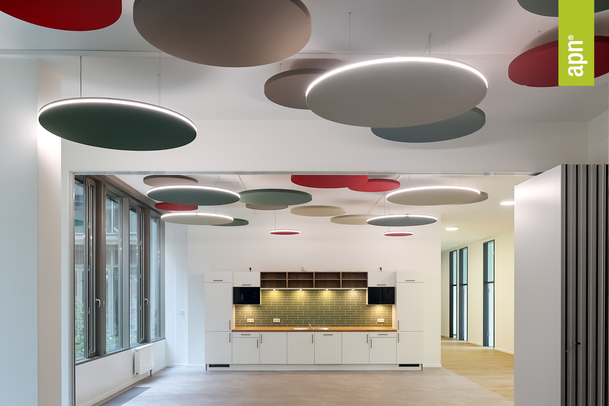 Moderner flexibler Raum mit Küche in minimalistischer Gestaltung, weißen Schränken und grüner Fliesenrückwand. An der Decke befinden sich runde Akustikpaneele in Rot, Grün und Braun mit integrierter Beleuchtung. Große Fenster auf der linken Seite lassen viel Tageslicht in den Raum.