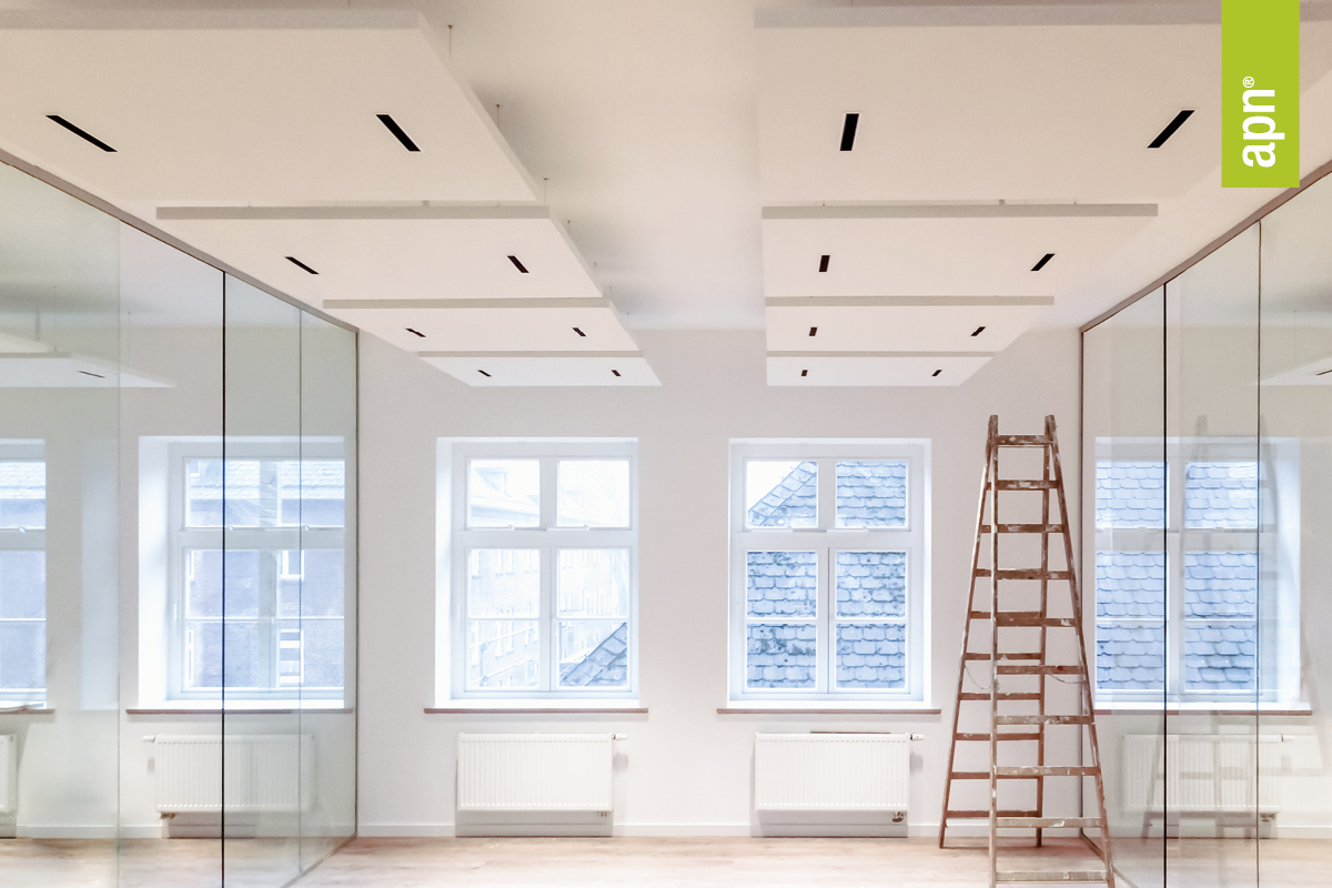 Minimalistisch gestalteter Raum mit großen Fenstern, Glaswänden und einer Holzleiter. An der Decke befinden sich rechteckige weiße PAkustikpaneele mit schwarzen Leuchten. Unter den Fenstern sind Heizkörper angebracht. Rechts im Bild ist ein grünes, vertikales Banner mit der Aufschrift „apn®“ zu sehen.