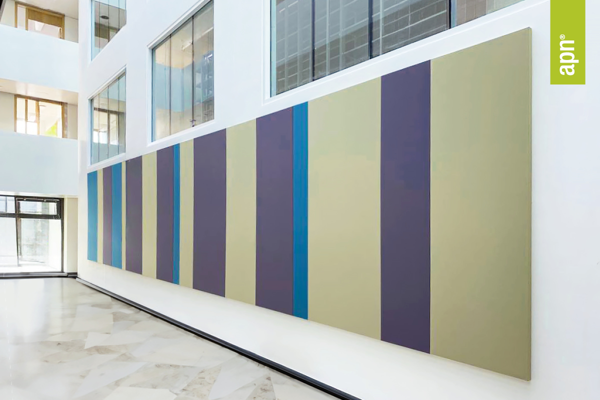 Moderner Innenraum mit großflächigem, abstraktem Wandkunstwerk aus vertikalen Farbstreifen in Grün-, Blau- und Lilatönen. Die Wandkunst besteht aus stoffbezogenen Akustikelementen. Große Fenster und weiße Wände sorgen für eine helle, offene Atmosphäre. Rechts im Bild befindet sich ein kleines grünes Banner mit der Aufschrift „apn®“.