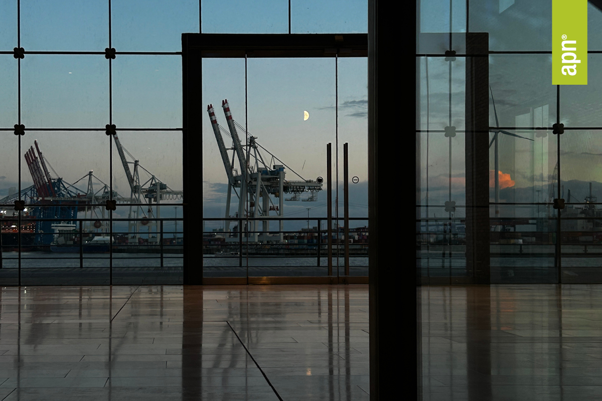 WELLENBRECHER: Das Bild zeigt den Blick aus einem Gebäude durch große Glasfenster auf einen industriellen Hafen. Mehrere Kräne sind in der Ferne zu sehen, silhouettiert gegen einen Dämmerungshimmel mit blauen und orangefarbenen Tönen am Horizont. Ein Halbmond ist über den Kränen sichtbar. Die Reflexion der Innenbeleuchtung ist auf dem Glas zu erkennen.