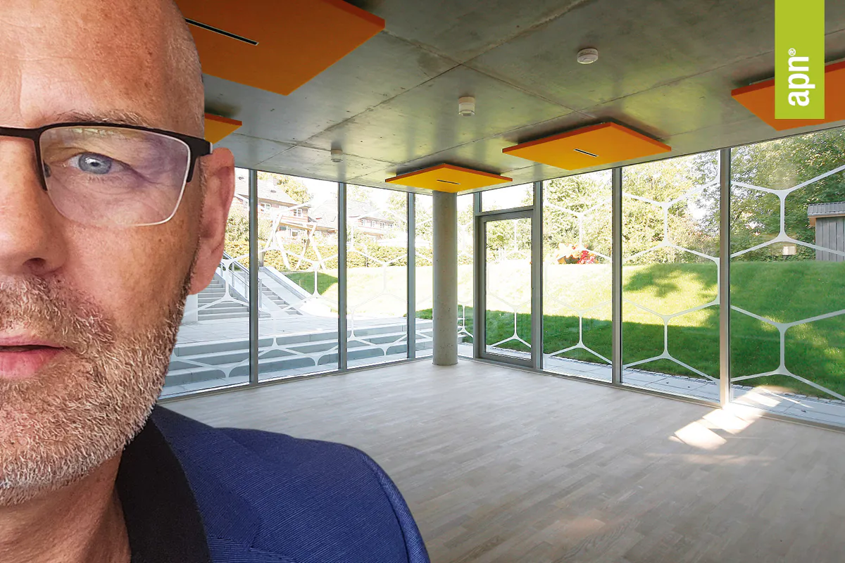 Leerer Raum mit großen Fensterfronten und Blick ins Grüne. Der Raum besteht aus Sichtbeton, an der Decke sind orange Lichtdeckensegel zu sehen. Im Vordergrund ist das halbe Profil eines mittelalten Mannes mit Brille, blauem Sakko und schwarzem Hemd zu sehen.