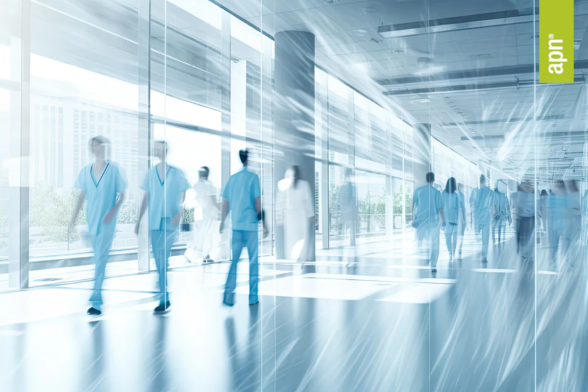 Das Bild zeigt eine moderne, helle Krankenhausumgebung mit mehreren Personen in blauen Kitteln, die durch einen Flur gehen.