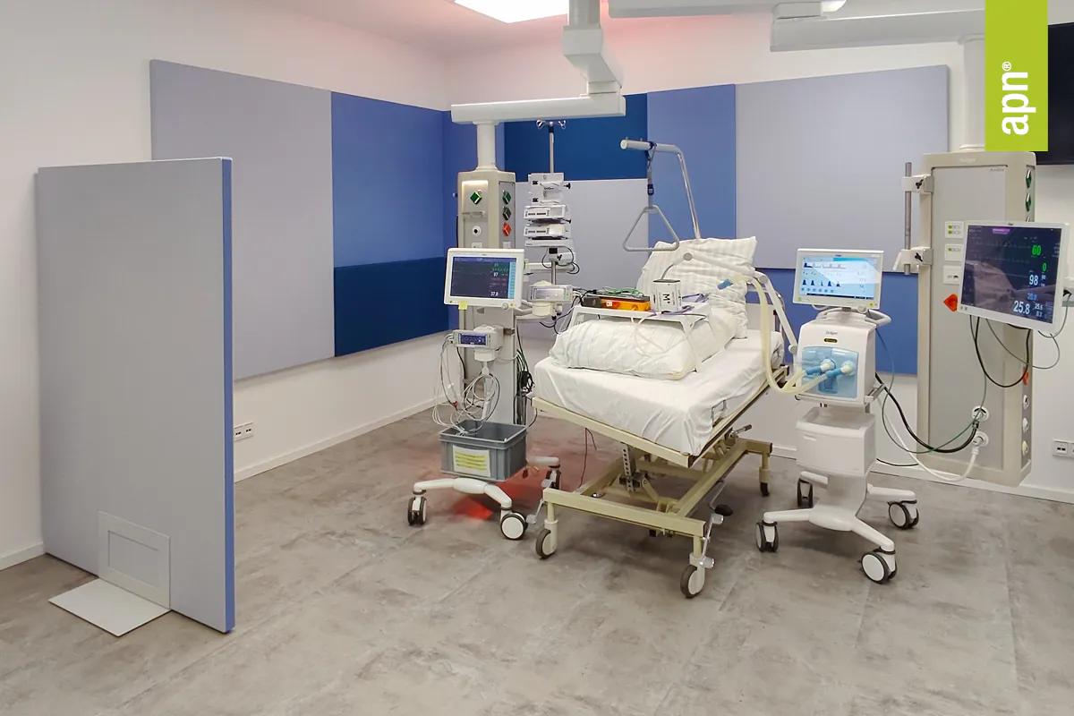 Ein modernes Krankenhauszimmer mit einem Krankenhausbett in der Mitte, umgeben von medizinischen Geräten und Monitoren. Das Bett ist mit weißen Kissen und Decken ausgestattet. Links vom Bett befindet sich ein Gerät auf einem Rollwagen mit einem Monitor darauf. Rechts vom Bett steht ein weiteres Gerät mit einem Bildschirm, das an eine Wandhalterung angeschlossen ist. Im Hintergrund sind blaue und graue Akustikpaneele an der Wand angebracht. Der Boden besteht aus grauem Fliesenbelag.