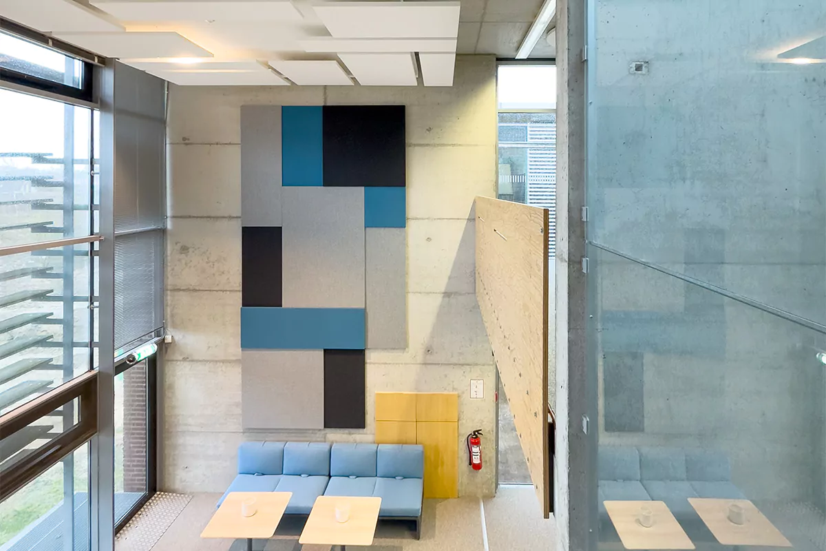 Innenraum einer modernen Hochschulbibliothek Gebäudes mit geometrischer akustischer Wandkunst in Blau- und Grautönen, davor ist ein hellblaues Sofa mit zwei Holztischen. Der Raum verfügt über große Fenster, die natürliches Licht hereinlassen.