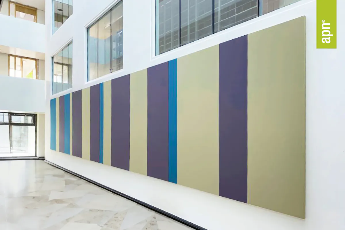 Moderner Innenraum mit großflächigem, abstraktem Wandkunstwerk aus vertikalen Farbstreifen in Grün-, Blau- und Lilatönen. Die Wandkunst besteht aus stoffbezogenen Akustikelementen. Große Fenster und weiße Wände sorgen für eine helle, offene Atmosphäre. Rechts im Bild befindet sich ein kleines grünes Banner mit der Aufschrift „apn®“.